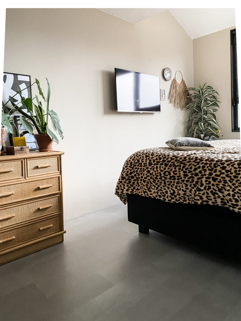 slaapkamer met betonlook laminaat van vtwonen, een beige wand, tijgerprint plaid over de zwarte boxspring en een vintage rotan kastje