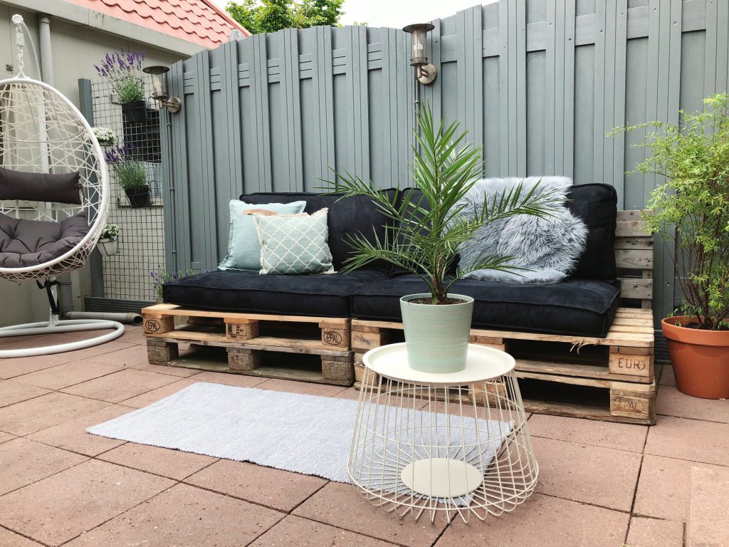 tuin make-over resultaat: dubbele pallet bank met zwarte kussens, metalen tafeltje met palm erop.